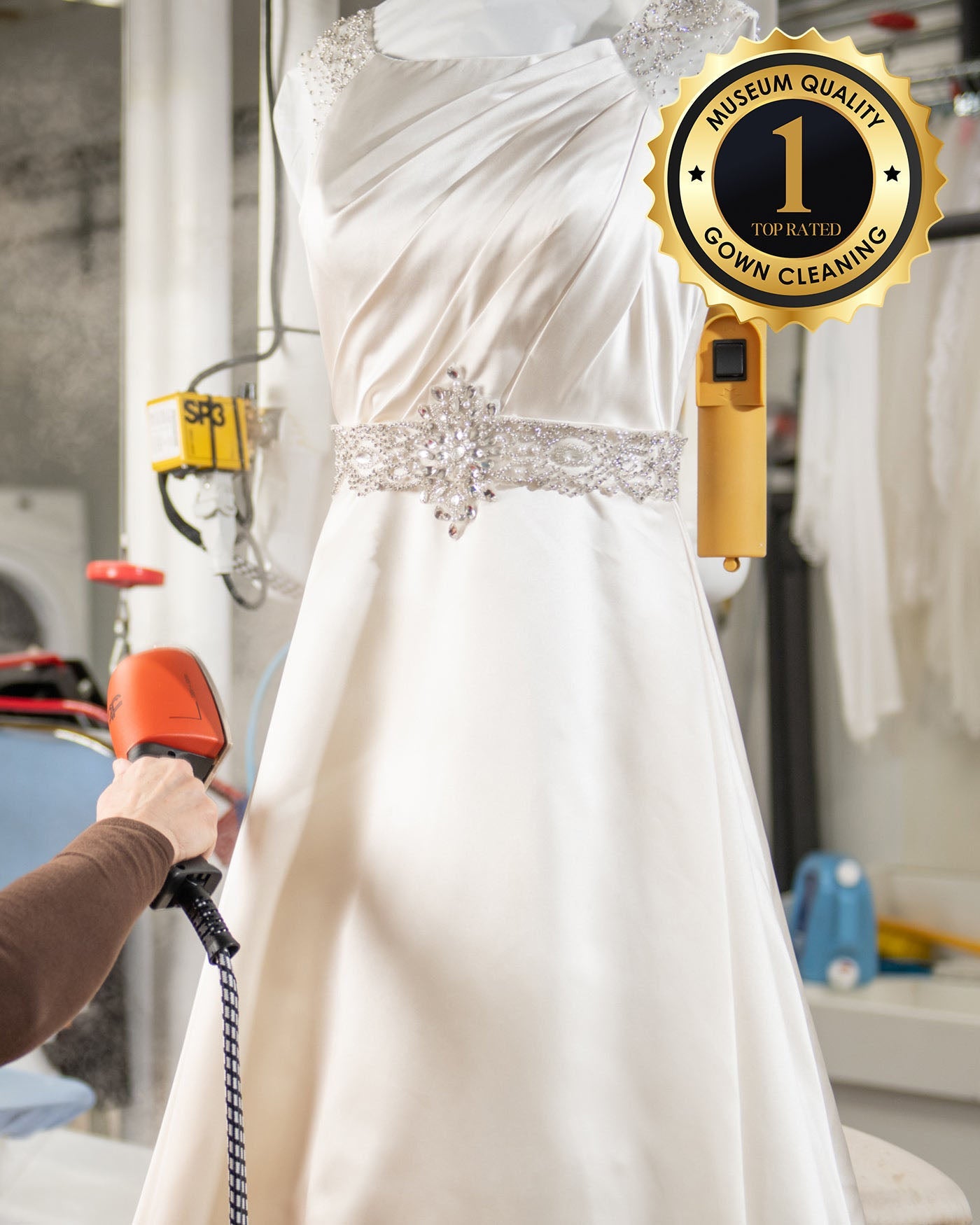 Using the Clean Only Wedding Dress Kit, someone steams a beaded white wedding gown displayed on a mannequin. A gold badge in the upper right corner says, “Museum Quality, Top Rated, #1 Gown Cleaning.”.