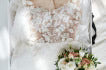 Close-up of STEP 4: Additional Items—a white lace wedding dress on a hanger, placed on a white surface with a bouquet of white and blush pink flowers and greenery beside it.